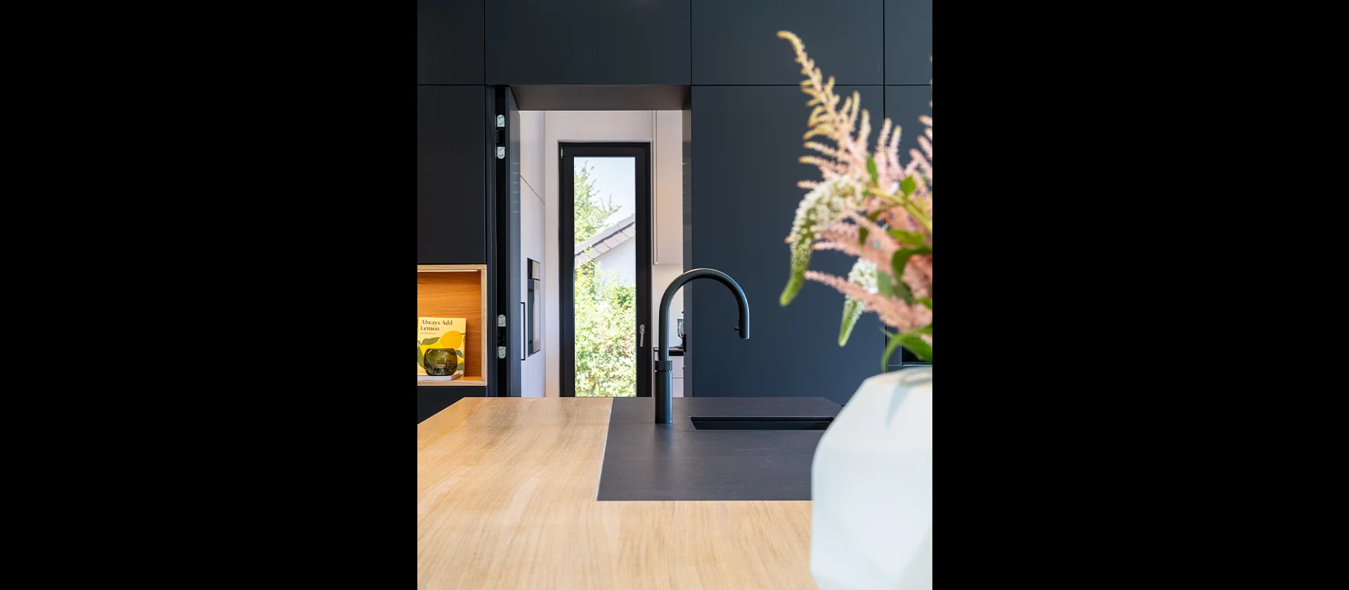 Detailaufnahme einer Kücheninsel aus hellem Holz mit schwarzer Spüle und Wasserhahn, im Hintergrund schwarze Schrankwand und Tür zum Garten