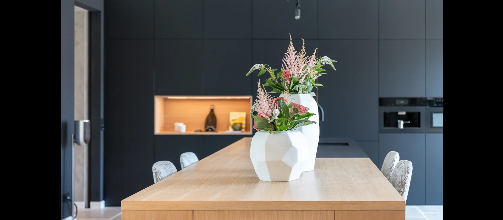 Nahaufnahme einer Kücheninsel aus Holz mit zwei weißen Vasen und frischen Blumen, im Hintergrund schwarze Küchenfront mit integriertem Backofen