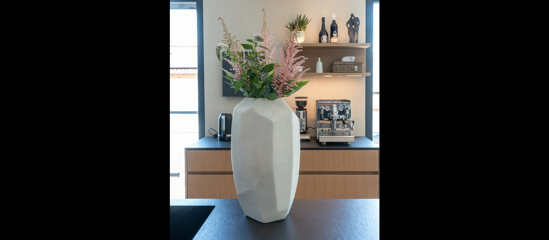 Detailansicht einer Küchenarbeitsfläche aus Fenix schwarz matt mit weißer Vase und Blumen, im Hintergrund Unterschränke aus Eiche und offene Regale mit Kaffeemaschine