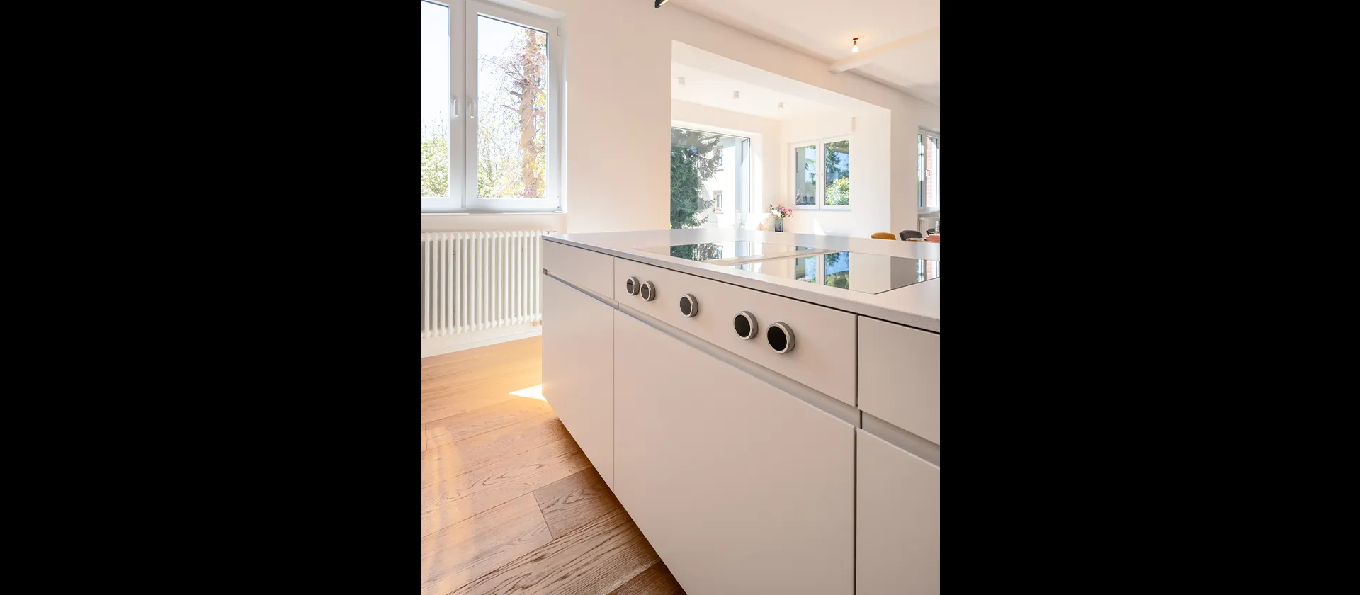 Detailansicht der Kochinsel mit eingelassenen Bedienknöpfen und Blick in den offenen Wohnbereich, präzise gefertigt von MTB Schreinerei.