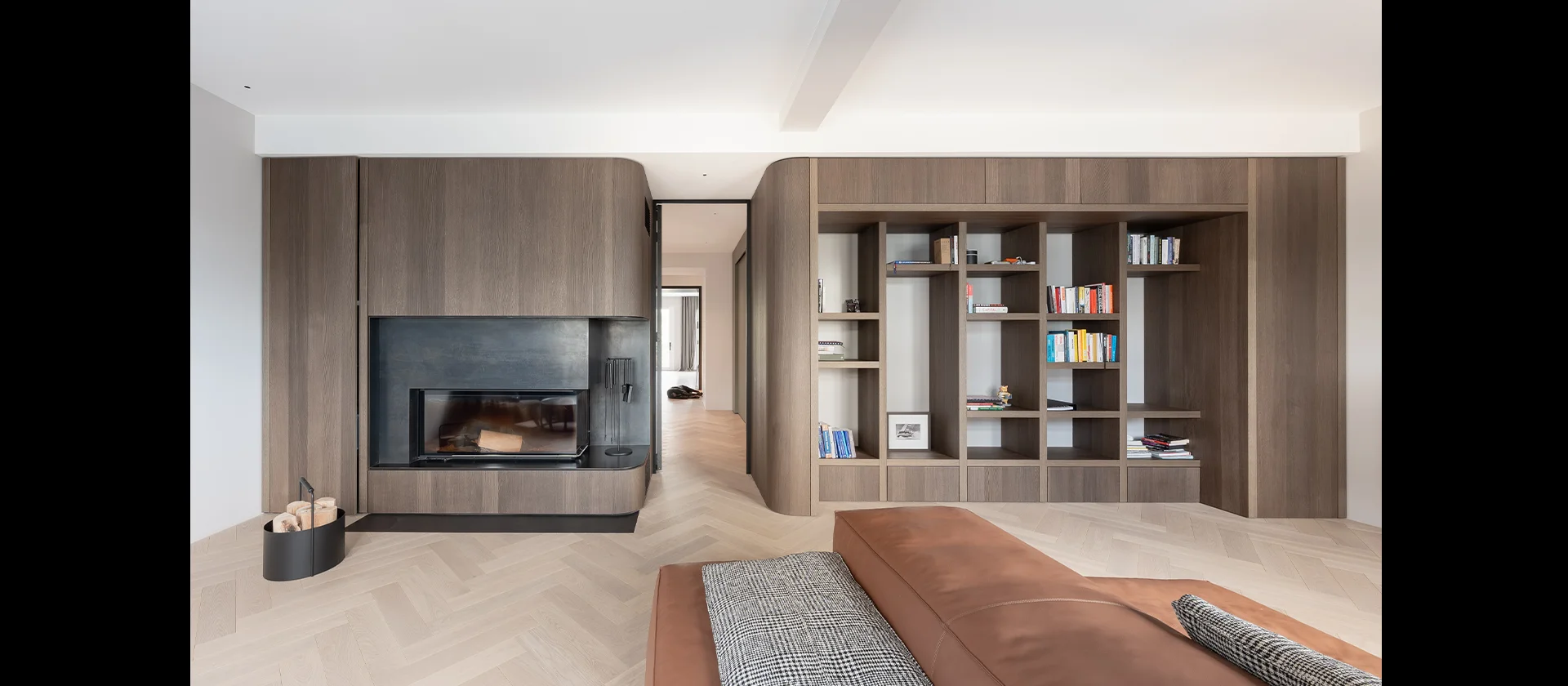 Weitläufiger Wohnraum mit eingelassenem Kamin und wandbündigem Bücherregal in dunkler Holzoptik, Blickrichtung in angrenzende Räume