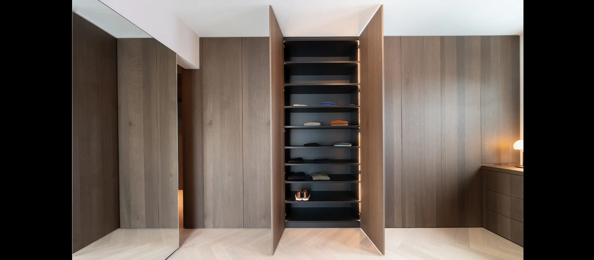 Offener Kleiderschrank mit dunkler Innenausstattung und integrierter Beleuchtung, eingebaut in eine wandbündige Holzverkleidung mit nahtlosen Türen