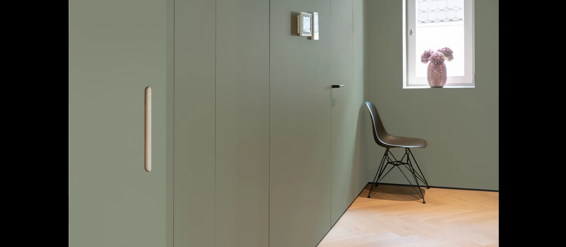 Grün lackierte, wandbündige Schrankanlage mit eingelassenen Griffleisten, daneben Fenster mit Deko-Vase und Designstuhl auf hellem Holzfußboden