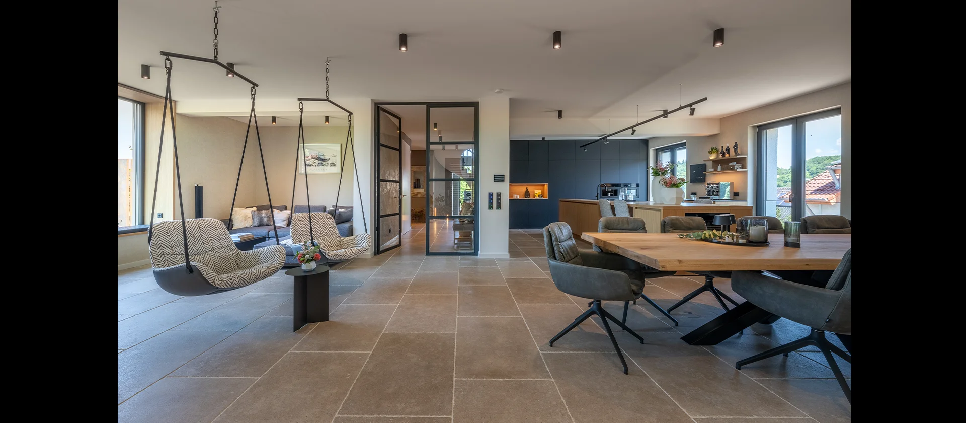 Offen gestalteter Wohn-Essbereich mit massivem Holztisch, gepolsterten Stühlen, Hängesesseln und moderner Einbauküche im Hintergrund