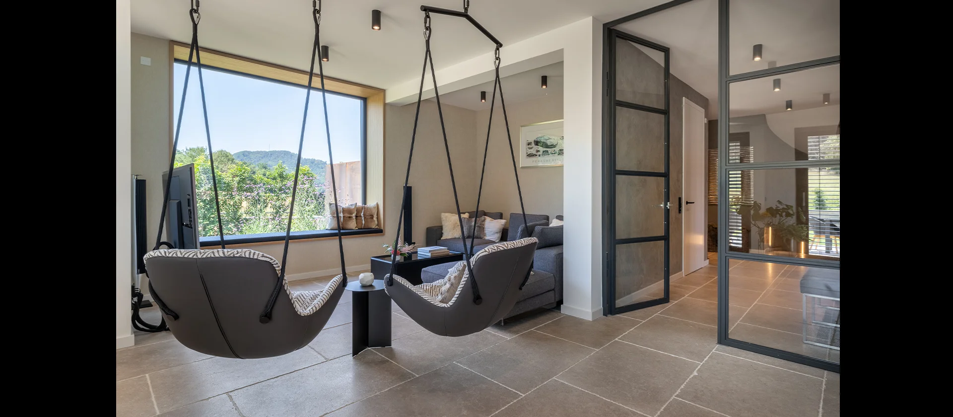 Gemütliche Wohnnische mit Hängesesseln, grauem Sofa, großem Fenster und Glastüren im modernen Metallrahmen
