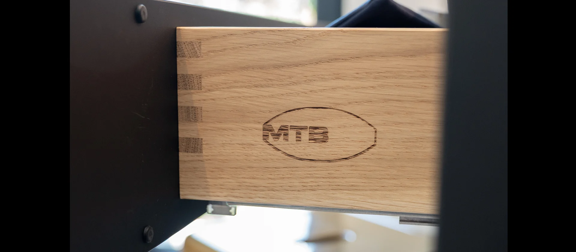 Detail einer massiven Holzschublade mit gezinkter Eckverbindung und eingebranntem MTB-Logo, eingebaut in einen dunklen Möbelkorpus mit Metallauszug