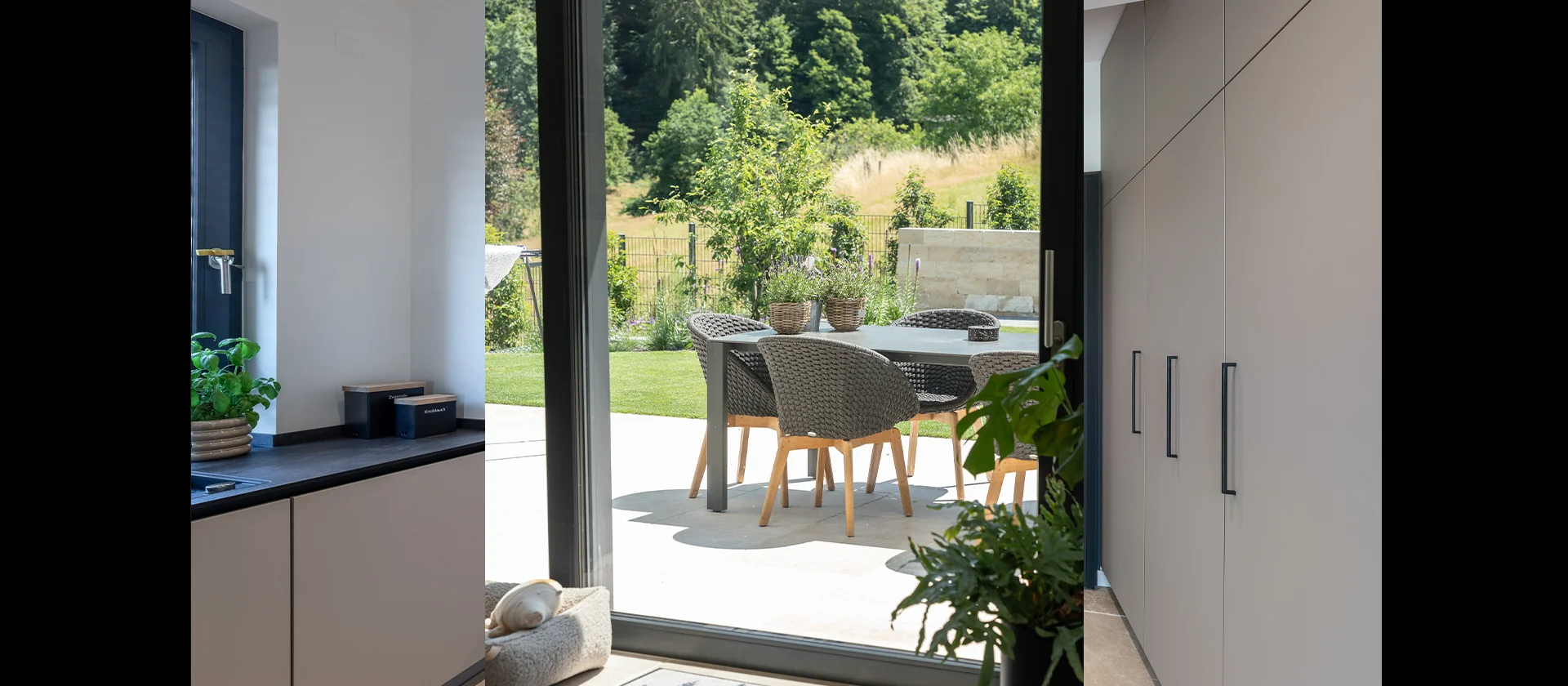 Blick von der Küche auf eine sonnige Terrasse mit Gartentisch und Rattanstühlen, bodentiefe Fenster und integrierter Pflanzbereich im Innenrau