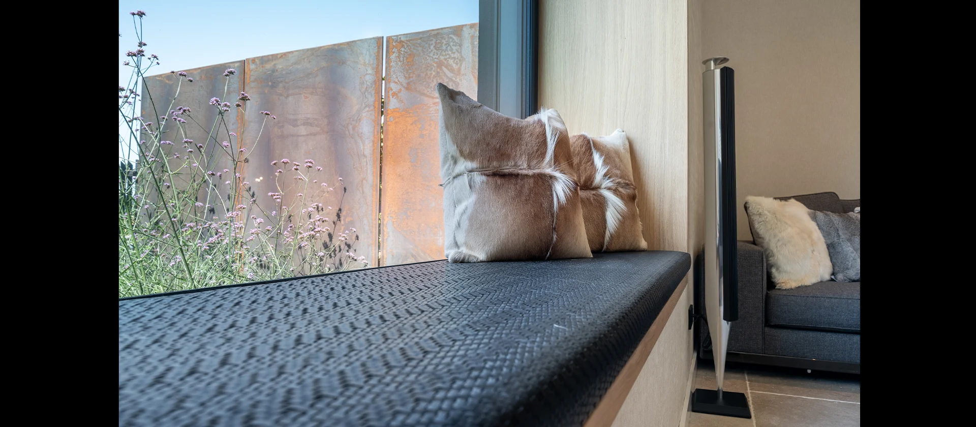 Fensterbank mit dunkler Sitzauflage und Kuhfellkissen vor großem Fenster, Blick nach draußen auf Gräser und rostfarbene Sichtschutzelemente